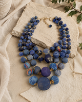 Blue four-strand kantha sari bead necklace laid flat on stone surface with linen fabric and green foliage, styled view