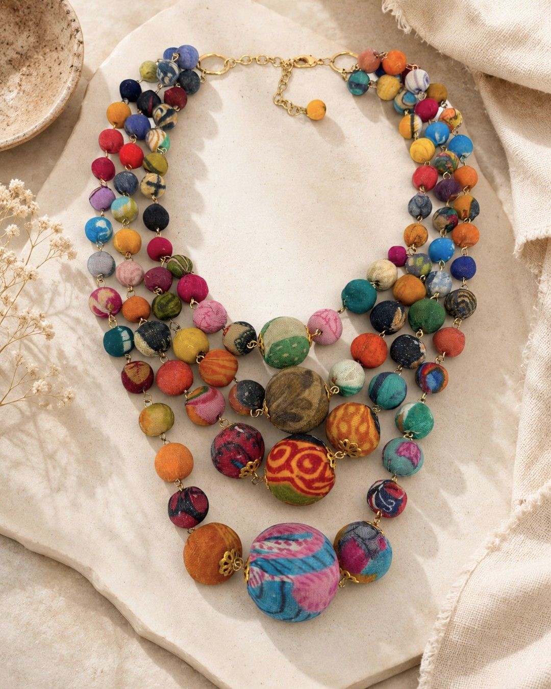 Kantha recycled textile necklace laid on linen fabric with a ceramic bowl and dried florals in the background