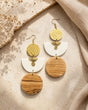 Kazuri brass and wood statement earrings laid flat on a textured stone surface alongside dried flowers and neutral linen fabric