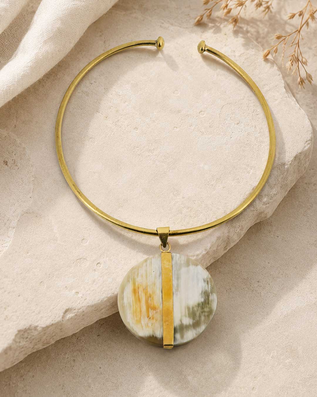 Brass collar choker with round horn disc pendant laid flat on a textured stone surface with dried grass in background, top-down view
