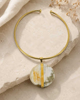 Brass collar choker with round horn disc pendant laid flat on a textured stone surface with dried grass in background, top-down view
