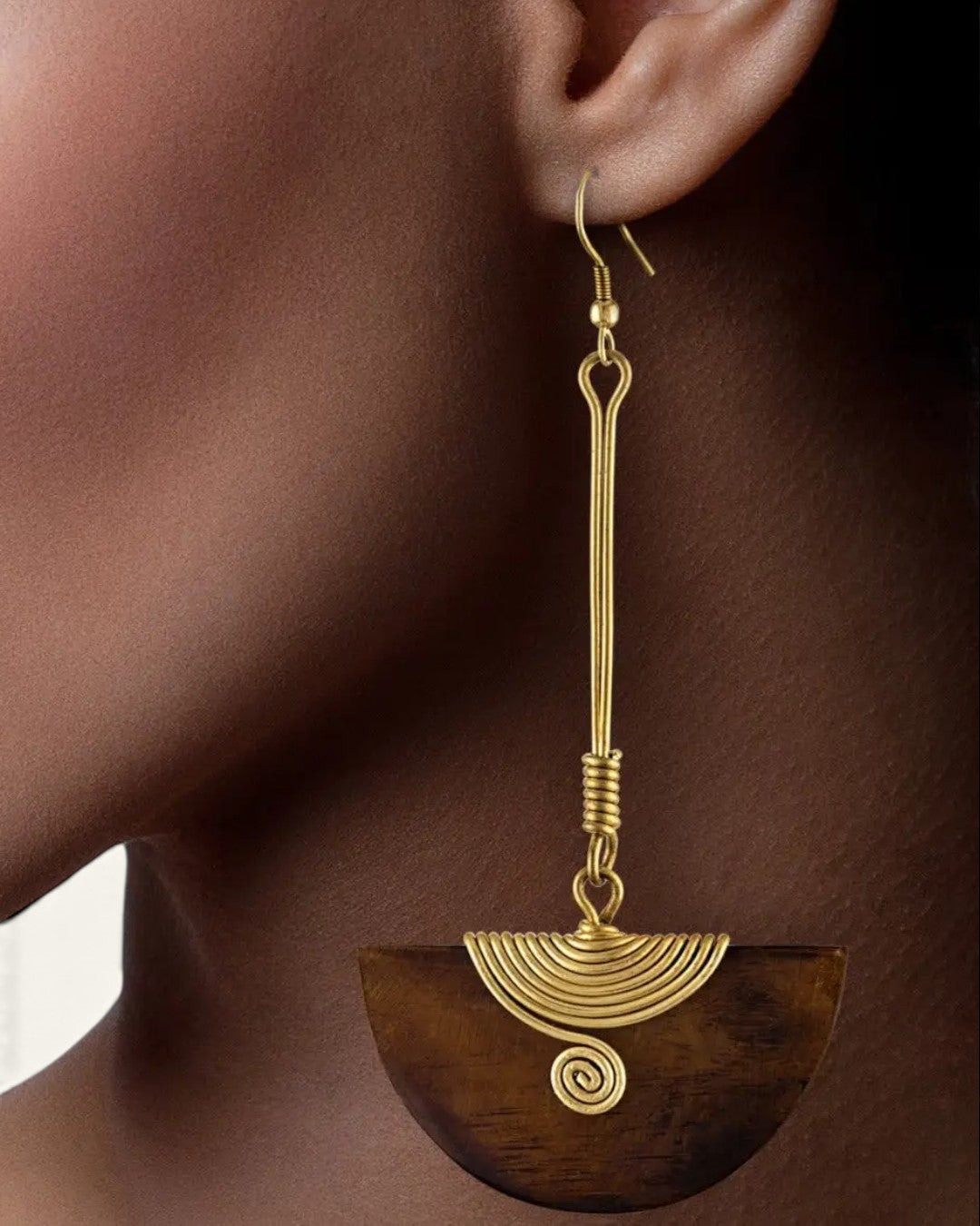 Close-up detail of one Kenyan brass wire spiral and dark wood half-moon pendant earring on model's ear against dark background