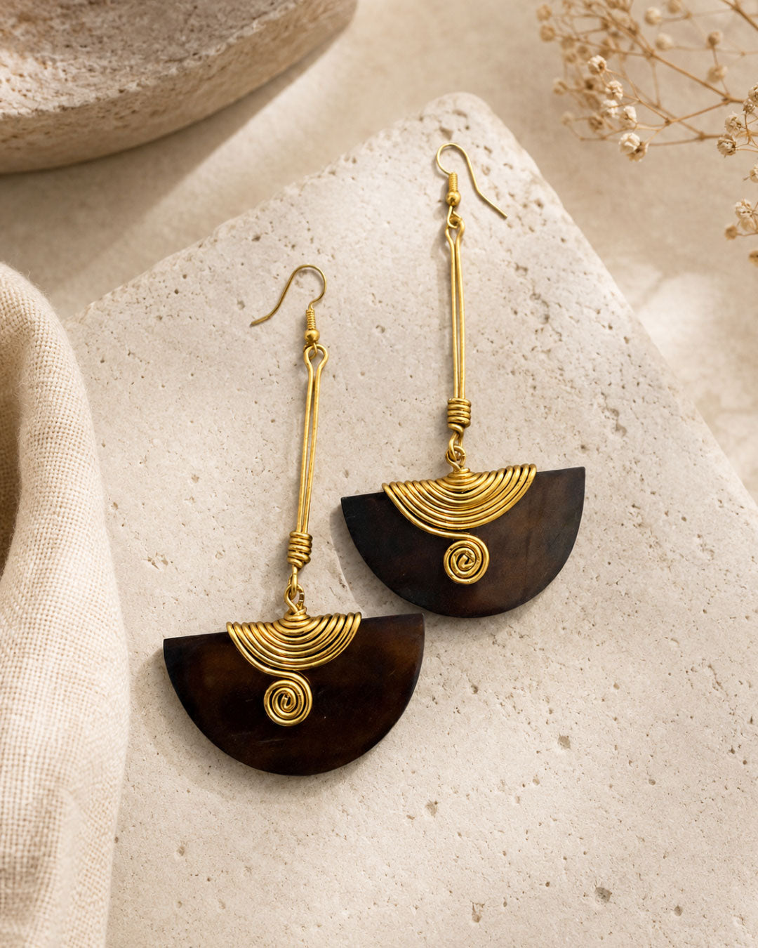 Brass and wood half-moon drop earrings styled on a stone surface with dried botanical stems and linen cloth in the background
