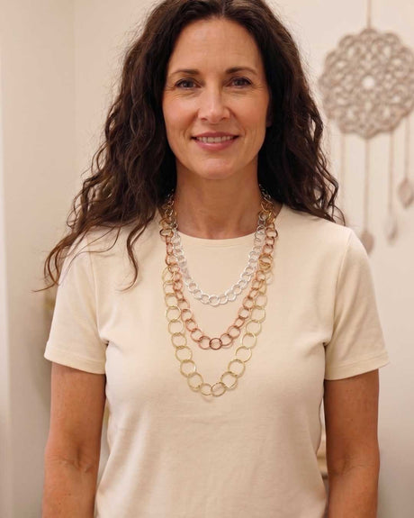 Three-strand mixed metal chain necklace worn by a woman in a cream t-shirt, showing layered gold, copper, and silver strands