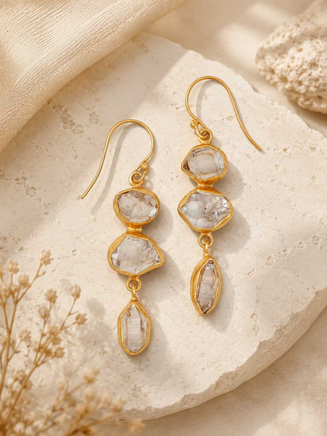Triple Herkimer quartz gold-plated drop earrings laid on a stone surface with dried flowers, styled lifestyle view