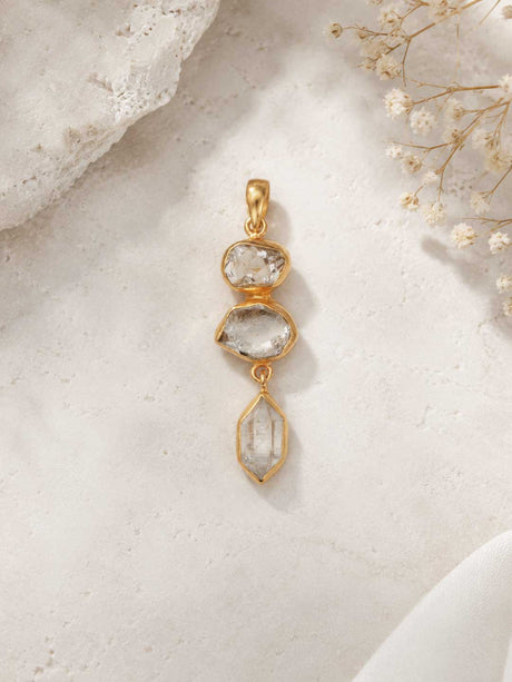 Triple Herkimer Quartz gold-plated pendant on a stone surface with dried flowers, showing three stacked crystals in gold bezels