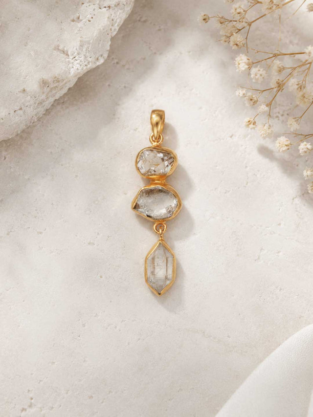 Triple Herkimer Quartz gold-plated pendant on a stone surface with dried flowers, showing three stacked crystals in gold bezels